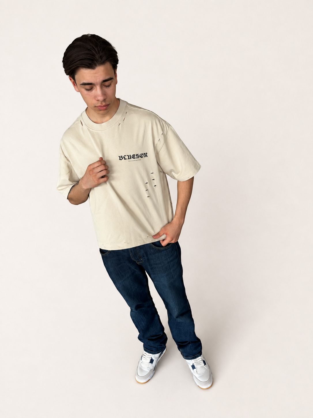 A young man wearing a cream-colored bluesor graphic t-shirt, dark blue jeans, and white sneakers, standing against a plain off-white studio background.