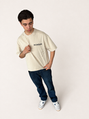 A young man wearing a cream-colored bluesor graphic t-shirt, dark blue jeans, and white sneakers, standing against a plain off-white studio background.