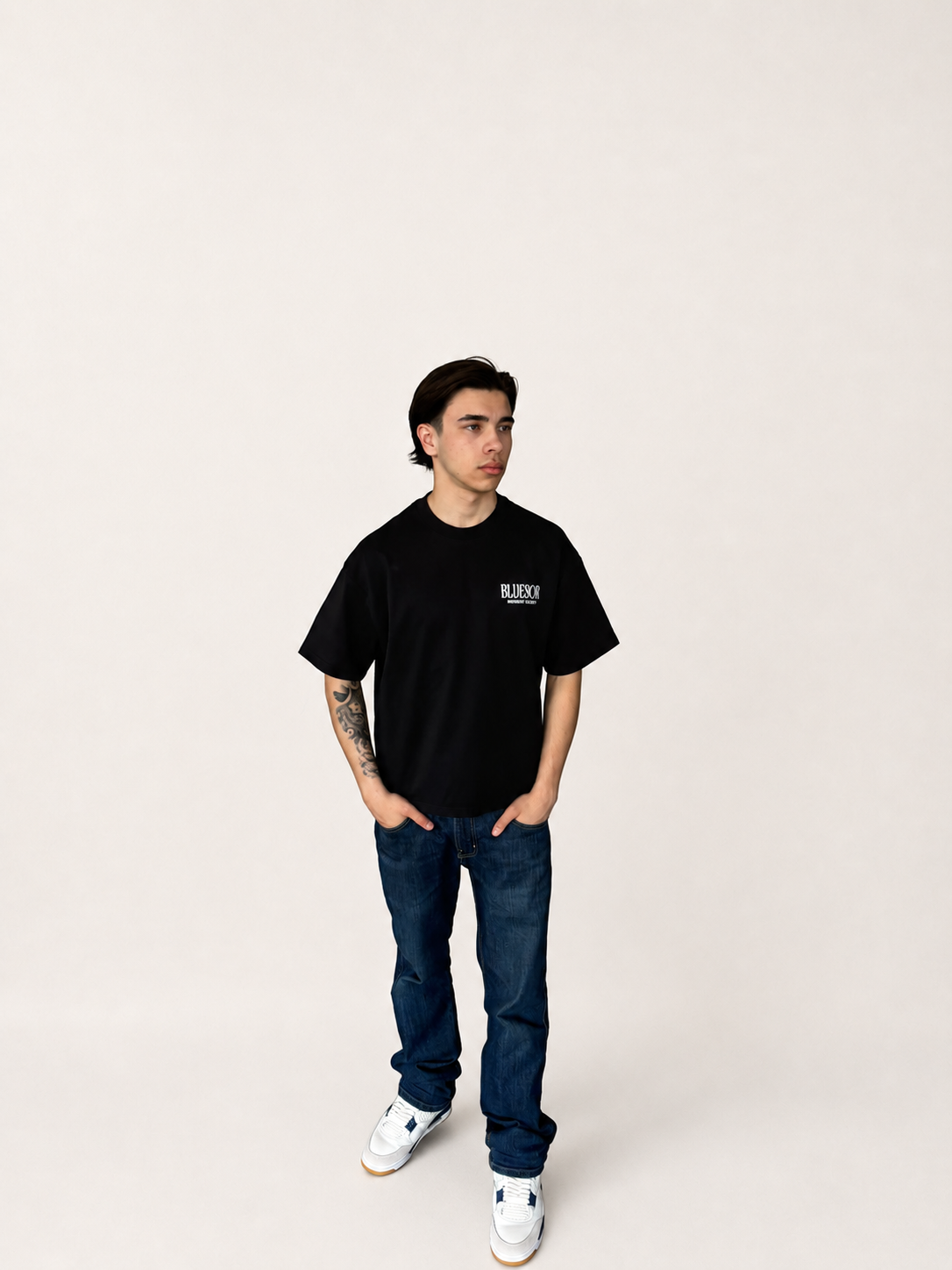 A man modeling a black short-sleeve t-shirt with a small white 'BLOOMY' logo on the left chest, paired with dark blue jeans and white sneakers, standing against a plain off-white studio background.