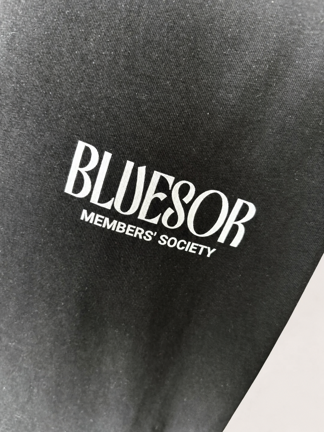 Close-up of a black fabric surface with white printed text reading 'BLUESOR' and smaller text 'MEMBERS' SOCIETY'.