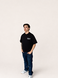 A man modeling a black crewneck t-shirt with a small white graphic on the left chest, paired with dark blue jeans and white sneakers, standing against a solid off-white background.