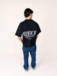 A man viewed from behind wearing a black oversized t-shirt with a large white 'BLUESOR MEMBERS SOCIETY' graphic on the back, paired with blue jeans and white sneakers, against a plain off-white studio background.