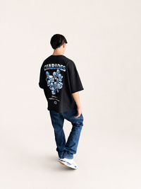 A man viewed from behind wearing an oversized black graphic T-shirt with a large blue and white skull print and the word 'CHARIDES' on the back, paired with blue jeans and white sneakers, against a plain light beige studio background.