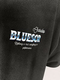 Close-up of a black T-shirt with a graphic print on the chest. The print features the word 'BLUESOR' in large blue and white gradient letters, with 'Charades' in white cursive above it and the phrase 'Nothing is real everything is performance' in smaller white script below.