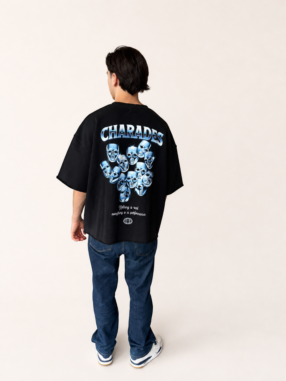 A person shown from the back wearing a black oversized T-shirt with a large blue and white graphic print and the word 'CHARADES' across the upper back, paired with dark blue jeans and white sneakers, against a plain light beige studio background.