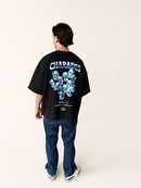 A person shown from the back wearing a black oversized T-shirt with a large blue and white graphic print and the word 'CHARADES' across the upper back, paired with dark blue jeans and white sneakers, against a plain light beige studio background.