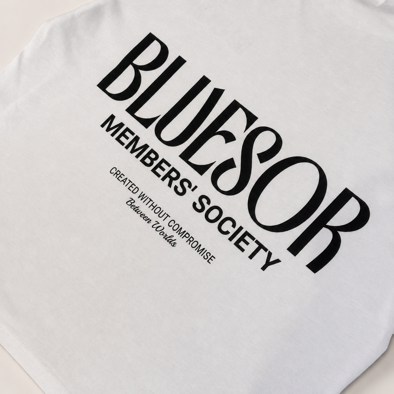 White t-shirt featuring a bold black graphic print on the front that reads 'BLUESOR MEMBERS' SOCIETY' with smaller text 'CREATED WITHOUT COMPROMISE Between Us'.