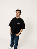 A man modeling an oversized black t-shirt with a white graphic print on the left chest, paired with blue jeans and white sneakers, standing against a plain off-white studio background.