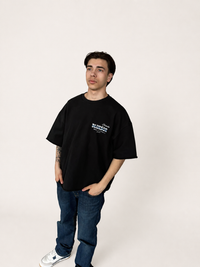 A man modeling an oversized black t-shirt with a white graphic print on the left chest, paired with blue jeans and white sneakers, standing against a plain off-white studio background.