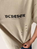 A close-up of a person wearing a beige oversized T-shirt with distressed holes and a black gothic 'BLUESOR' graphic across the chest, with 'TRANSCENDENT' printed underneath.