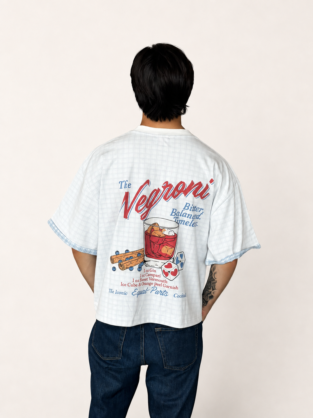 A man viewed from behind wearing a white short-sleeve graphic t-shirt with a light blue gingham pattern and a large retro-style 'The Negroni' cocktail print on the back, paired with dark blue jeans against a plain off-white background.