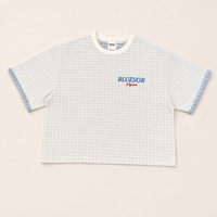 A short-sleeve crop top t-shirt with a white and light blue gingham pattern, featuring a white crew neck, blue and red 'BLUESOR Megami' text on the chest, and blue patterned sleeve cuffs, laid flat on a light beige background.