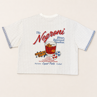 A white short-sleeve t-shirt with a light blue gingham pattern, featuring a retro-style 'The Negroni' cocktail graphic on the front. The graphic includes red and blue text, a glass of red drink, and illustrations of a cinnamon stick and blueberries.