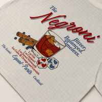 A close-up of a light beige or off-white fabric surface featuring a printed graphic for 'The Negroni Cocktail'. The graphic includes red and blue text, an illustration of a glass with red liquid and ice, blue gin balls, cinnamon sticks, and heart-patterned dice.