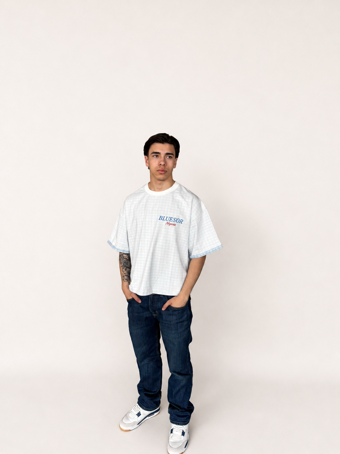 A man modeling an oversized white gingham t-shirt with blue and red 'BILLYRUSH' text on the chest, paired with dark blue jeans and white sneakers, standing against a plain off-white background.
