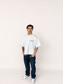 A man modeling an oversized white gingham t-shirt with blue and red 'BILLYRUSH' text on the chest, paired with dark blue jeans and white sneakers, standing against a plain off-white background.