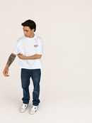 A man wearing a white short-sleeve t-shirt with a small black graphic on the left chest, paired with dark blue jeans and white sneakers, standing against a solid off-white background.