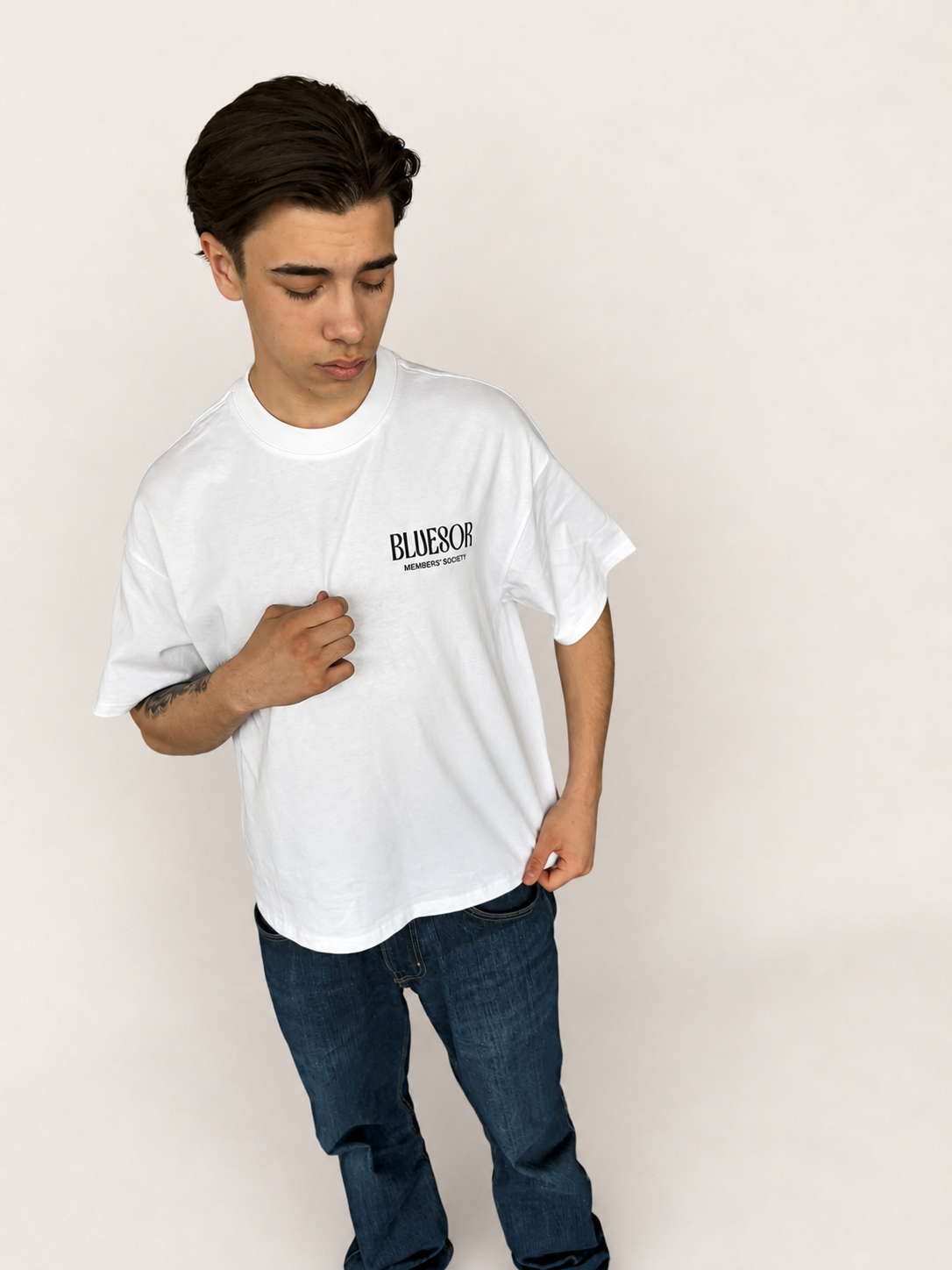 A young man wearing an oversized white t-shirt with a black 'BLAZERSPORT' logo on the chest, paired with dark blue jeans, standing against a plain off-white studio background.