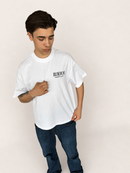 A young man wearing an oversized white t-shirt with a black 'BLAZERSPORT' logo on the chest, paired with dark blue jeans, standing against a plain off-white studio background.