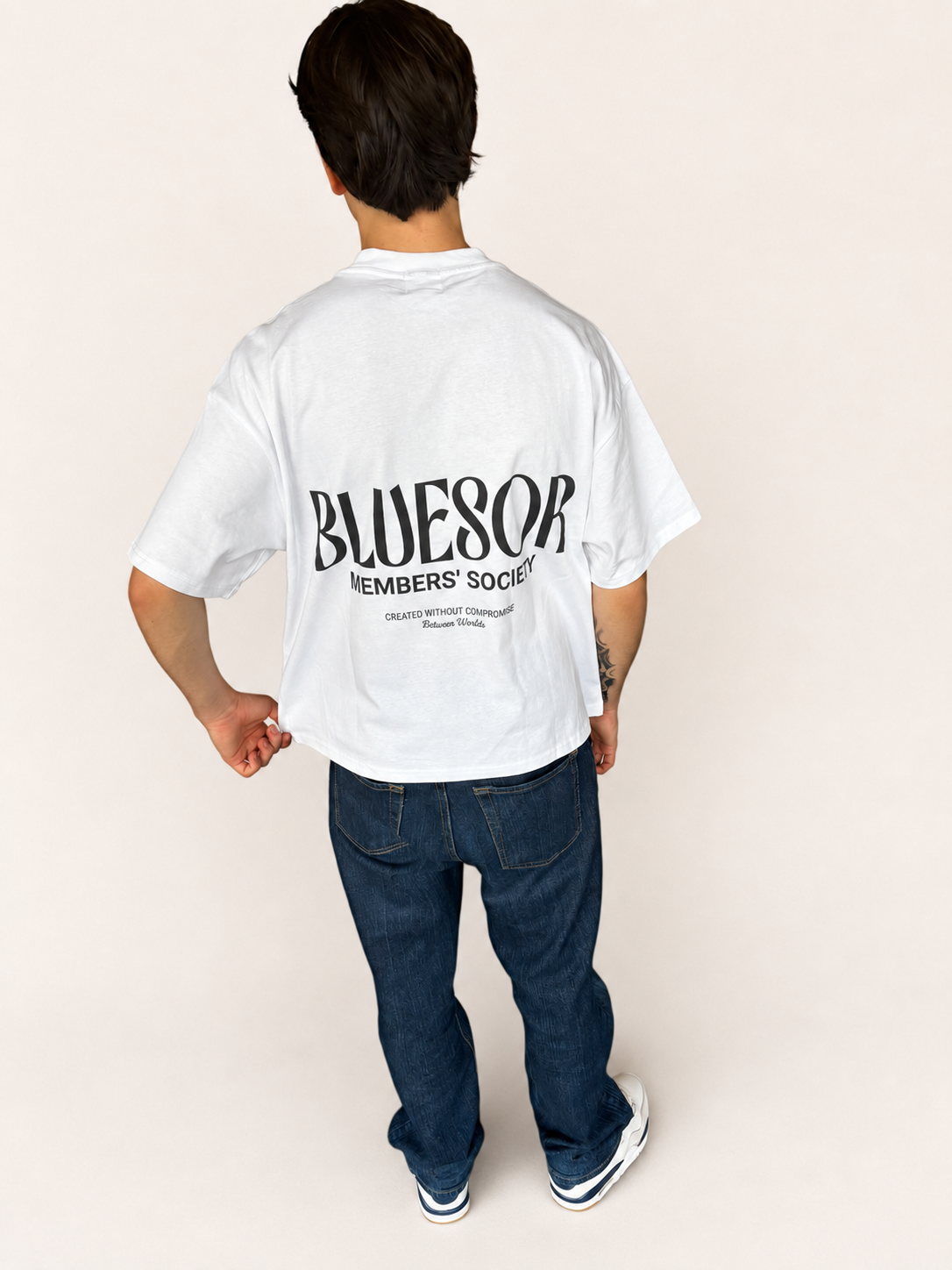 A person shown from the back wearing an oversized white t-shirt with a large black 'BLUESOR MEMBERS' SOCIETY' graphic printed on the back, paired with dark blue jeans and white sneakers, against a solid light beige background.