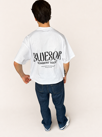 A person shown from the back wearing an oversized white t-shirt with a large black 'BLUESOR MEMBERS' SOCIETY' graphic printed on the back, paired with dark blue jeans and white sneakers, against a solid light beige background.